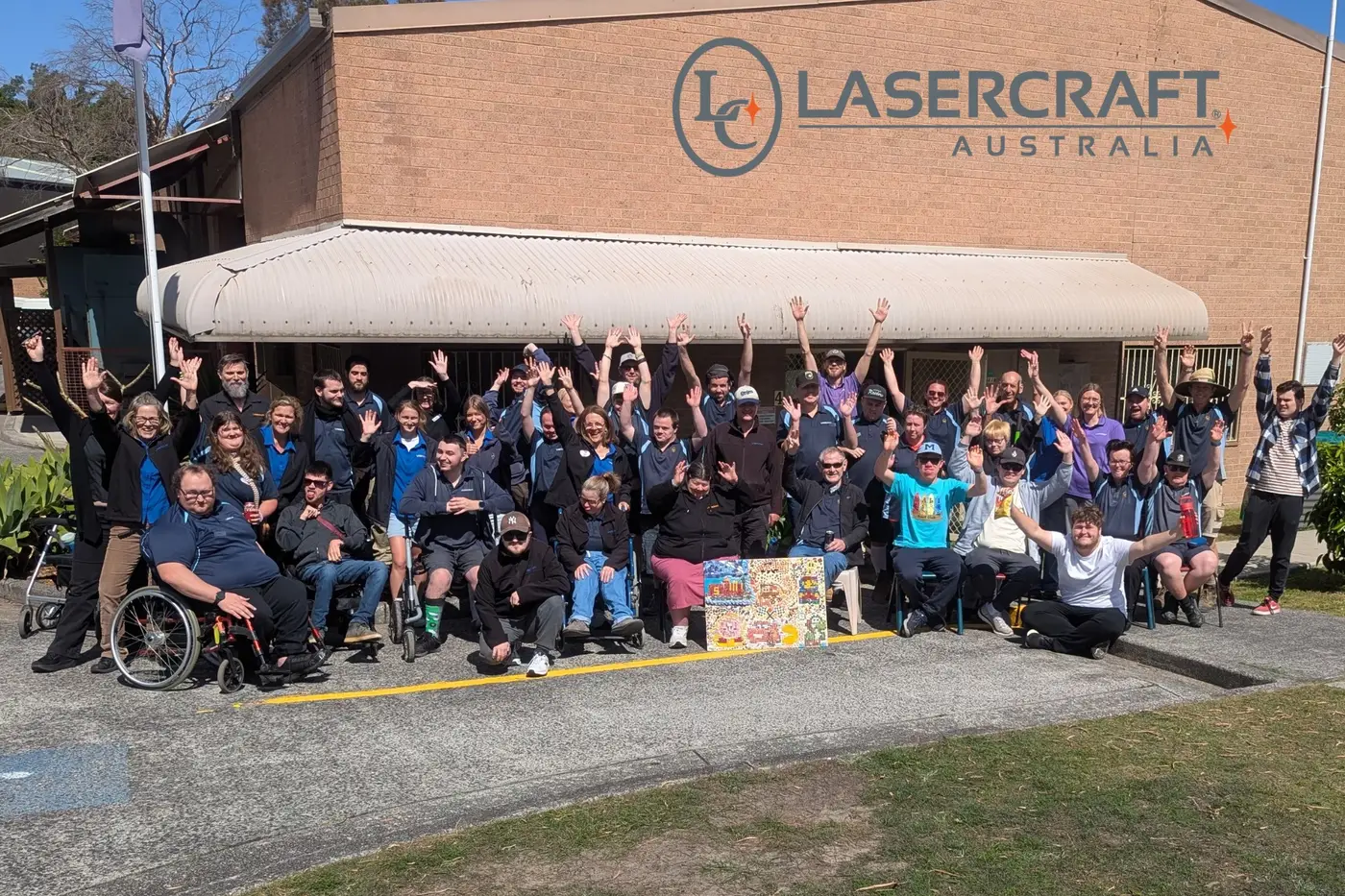 The whole team gathered outside the West Gosford building under the Lasercraft Australia sign, most with their arms raised.