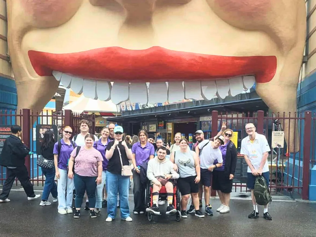 A group in purple shirts standing together on the footpath under the big smiling face of the Luna Park entrance in Sydney.