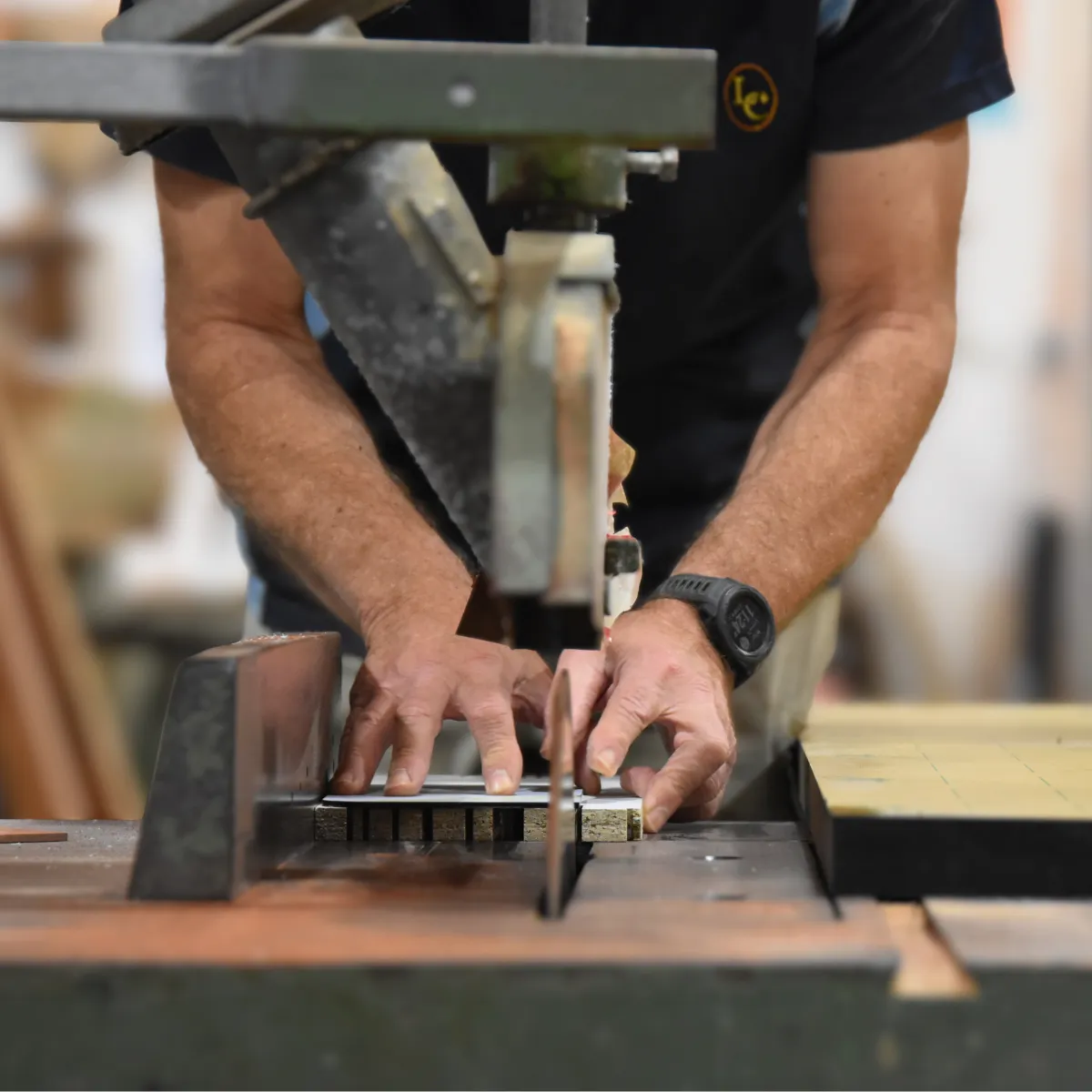Two hands guiding a stack of timber blanks through a saw on the workshop bench, the Lasercraft mark on the operator's shirt.