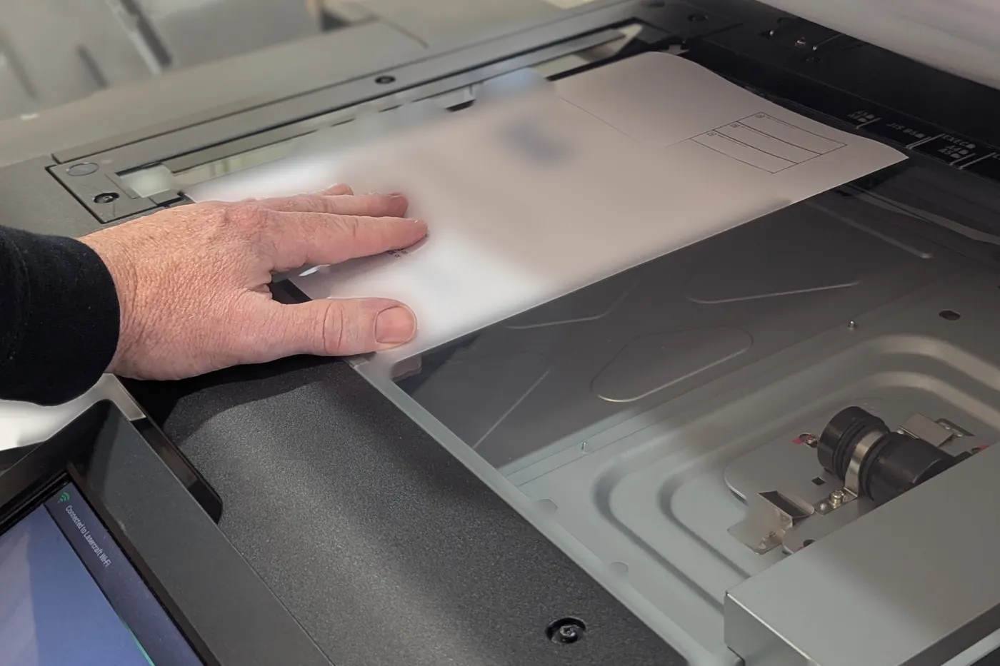 A hand holding a sheet of paper flat on the glass of an office scanner, ready to scan.
