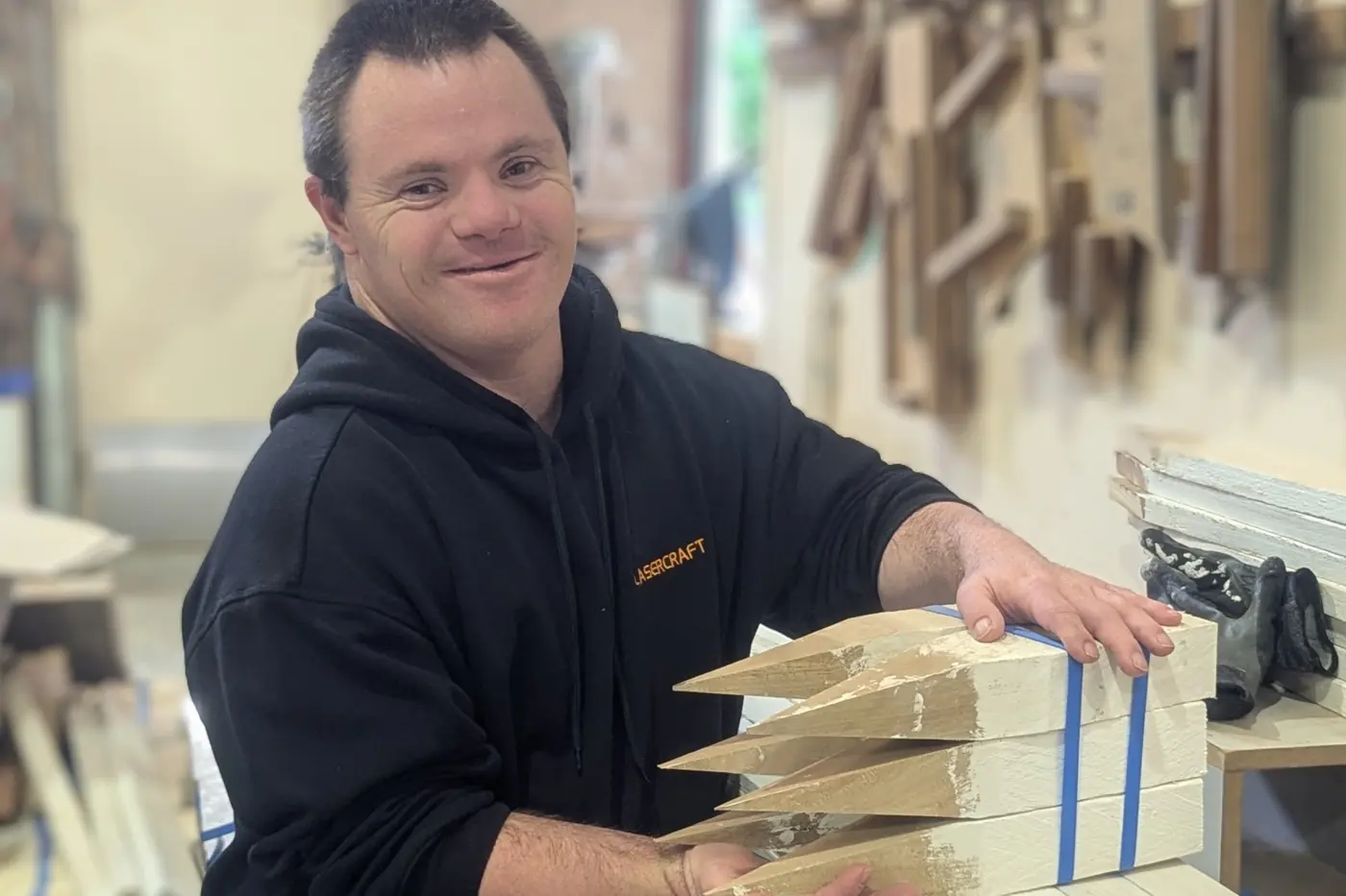 A Lasercraft employee in a black hooded jumper carrying the company name in orange, standing at a bench in the West Gosford workshop with two bundles of white painted timber survey pegs resting across both hands, each peg sharpened to a point and each bundle strapped with blue banding, and offcuts of timber hanging on the wall behind.